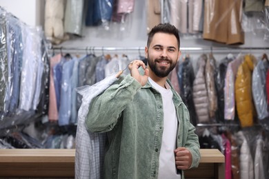 Photo of Dry-cleaning service. Happy man holding hanger with shirt in plastic bag indoors