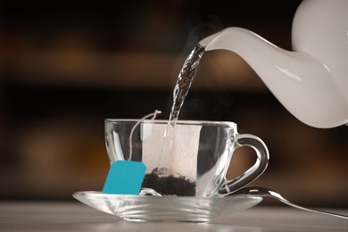 Pouring hot water into glass cup with tea bag on table against blurred background