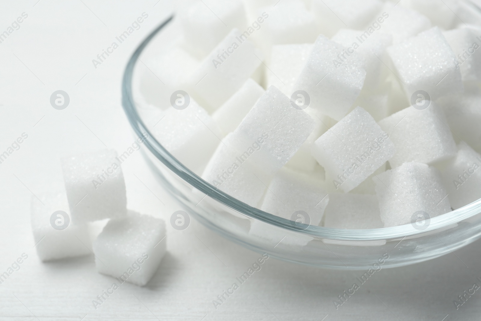 Photo of Many sugar cubes in glass bowl on white wooden table, closeup