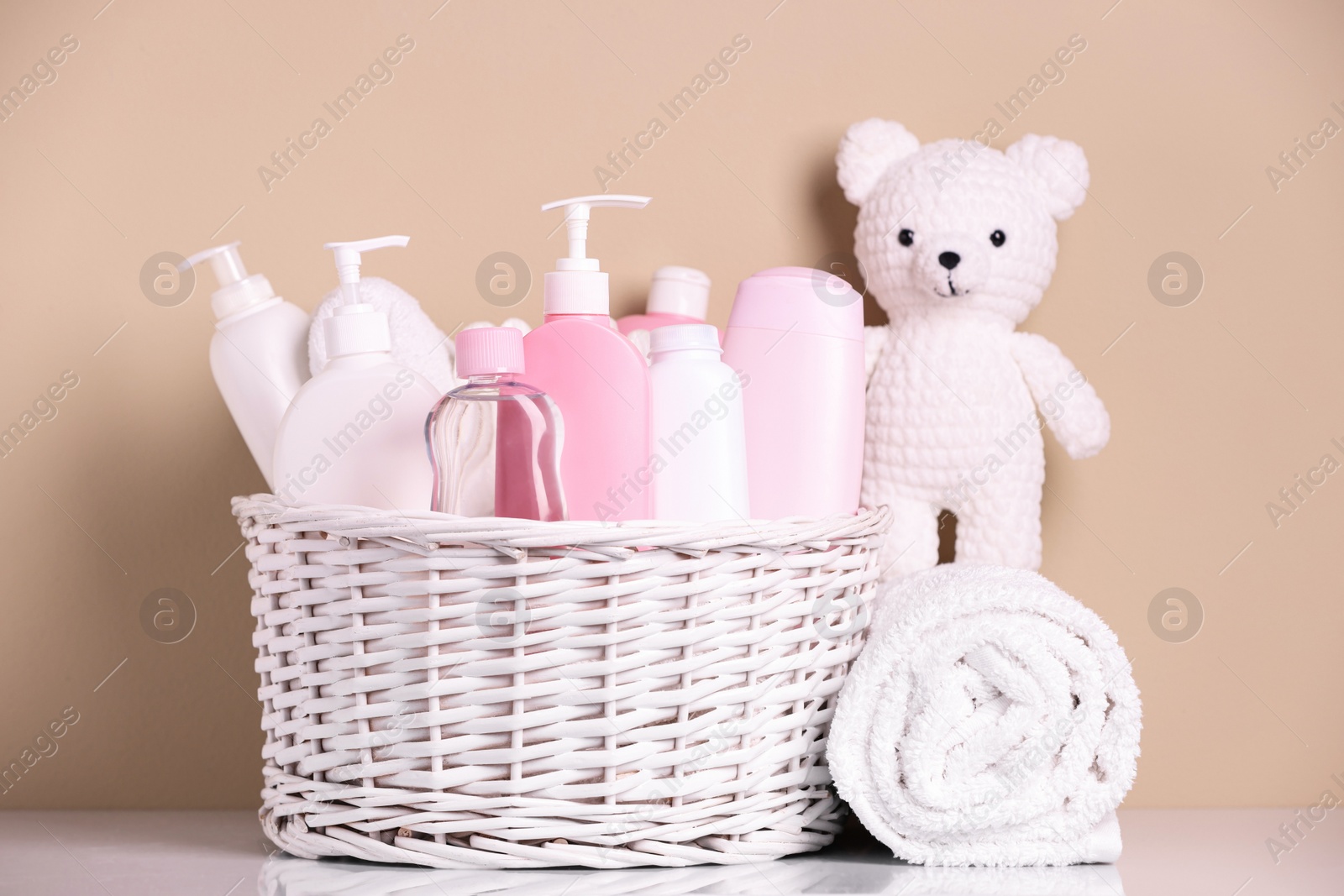 Photo of Wicker basket with baby cosmetic products, bath accessories and knitted toy bear on white table against beige background