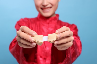 Photo of Young woman holding tasty fortune cookie with prediction on light blue background, closeup