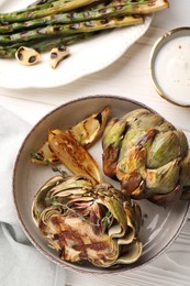 Tasty grilled artichokes served with sauce and asparagus on white wooden table, flat lay