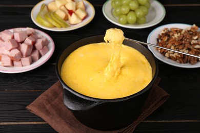 Dipping piece of ham into fondue pot with melted cheese on black wooden table, closeup