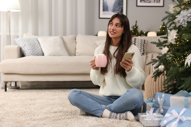 Photo of Smiling woman with cup of Christmas cocoa and smartphone at home. Space for text