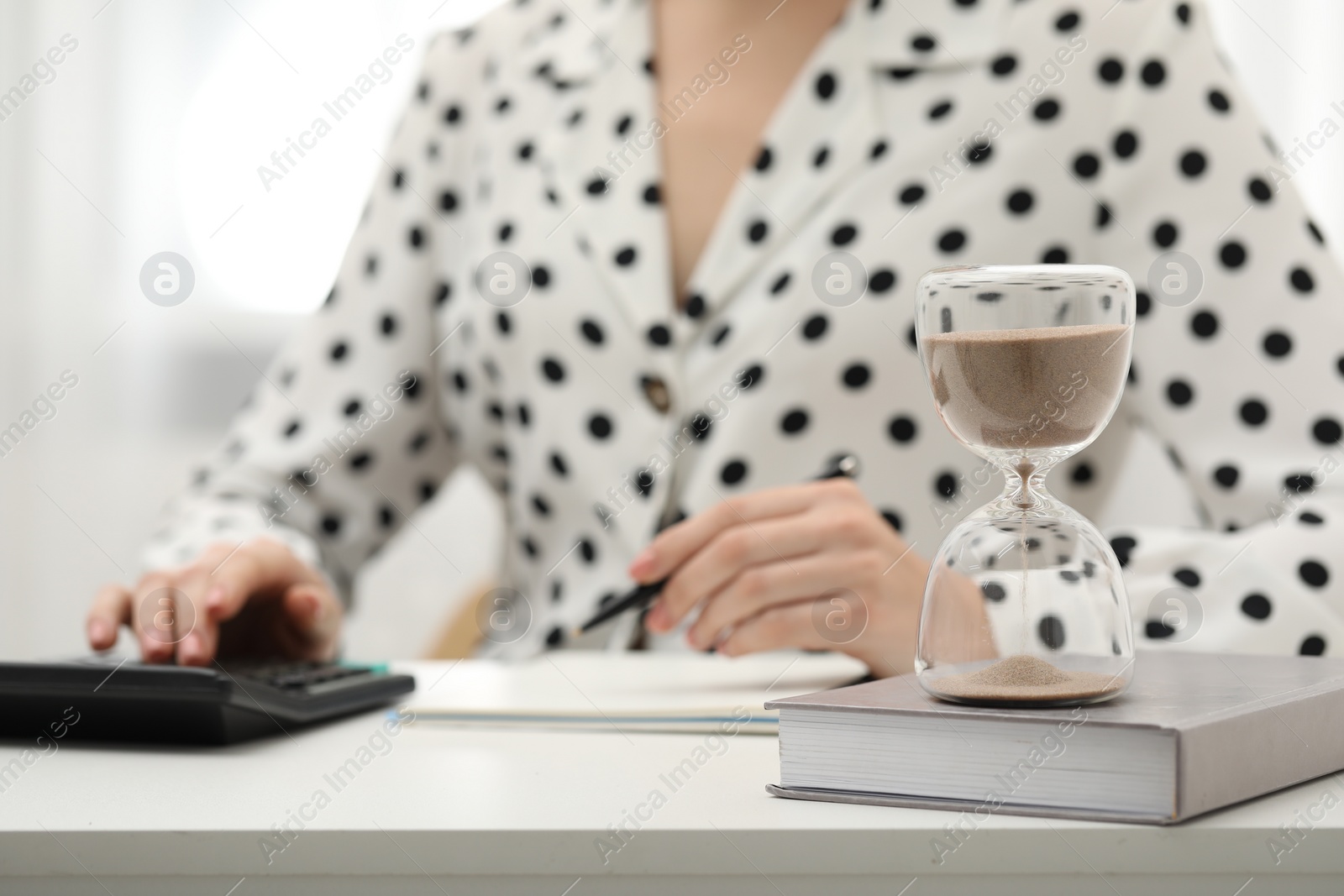 Photo of Hourglass with flowing sand on desk. Woman taking notes while using calculator indoors, selective focus