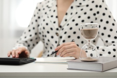 Hourglass with flowing sand on desk. Woman taking notes while using calculator indoors, selective focus