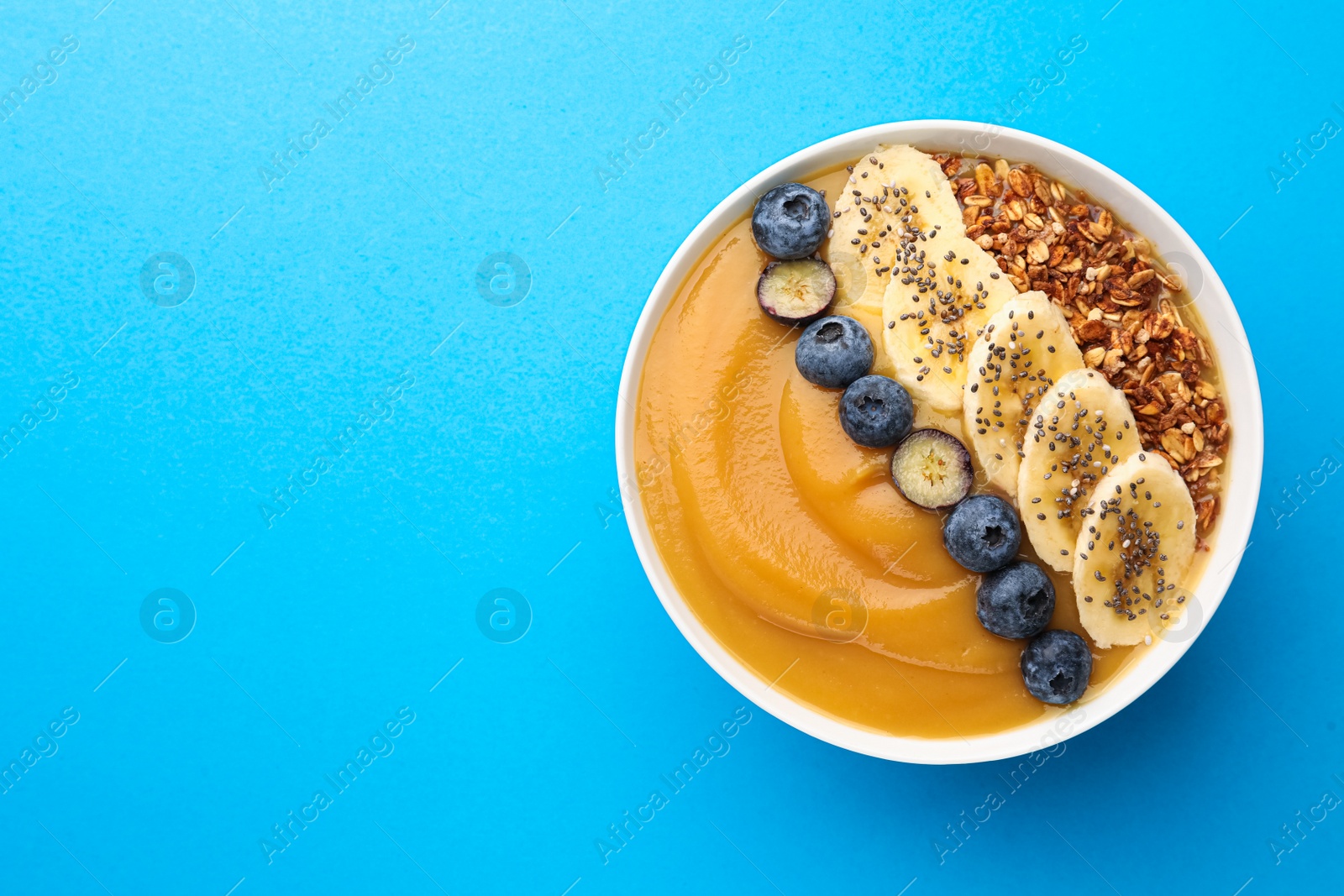 Photo of Delicious smoothie bowl with fresh blueberries, banana and granola on light blue background, top view. Space for text