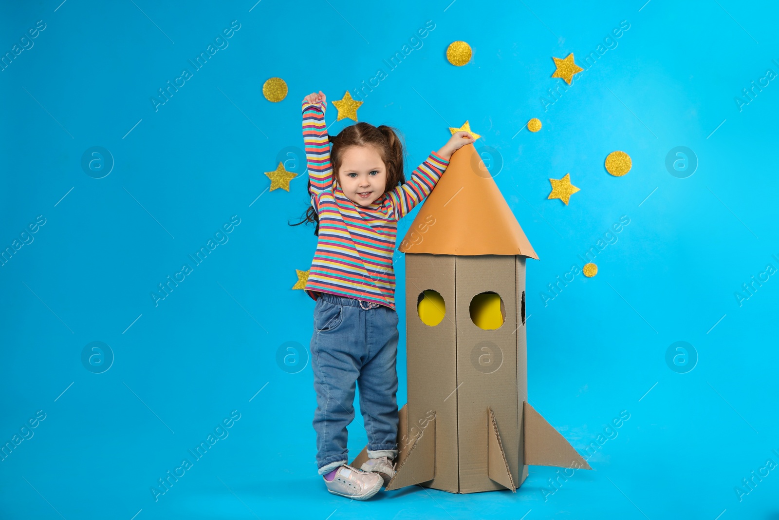Photo of Little child playing with rocket made of cardboard box near stars on blue background
