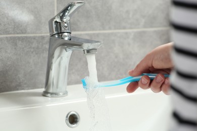 Woman washing plastic toothbrush under flowing water from faucet in bathroom, closeup