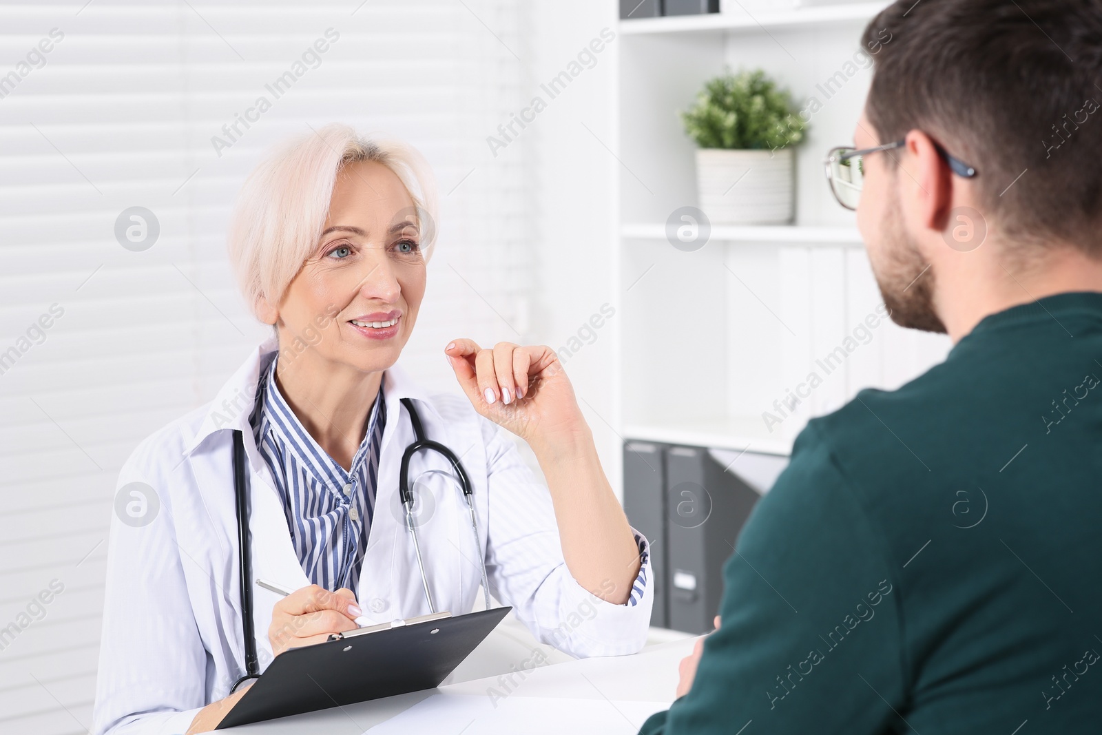 Photo of Doctor with pen and clipboard consulting patient at table in clinic