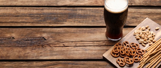 Glass of beer served with delicious pretzel crackers and other snacks on wooden table, space for text. Banner design