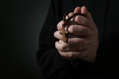 Photo of Priest with rosary beads praying on dark background, closeup. Space for text