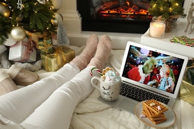 Photo of MYKOLAIV, UKRAINE - DECEMBER 23, 2020: Woman watching The Grinch movie on laptop near fireplace at home, closeup. Cozy winter holidays atmosphere