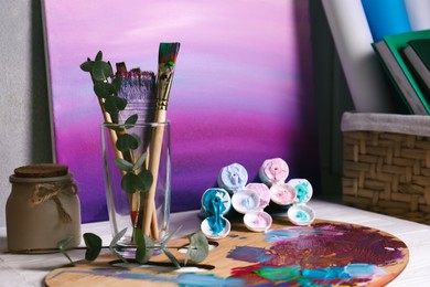 Wooden artist's palette with colorful paints, brushes and scented candle on white table