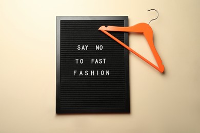 Photo of Black letter board with phrase SAY NO TO FAST FASHION, orange hanger on beige background, flat lay