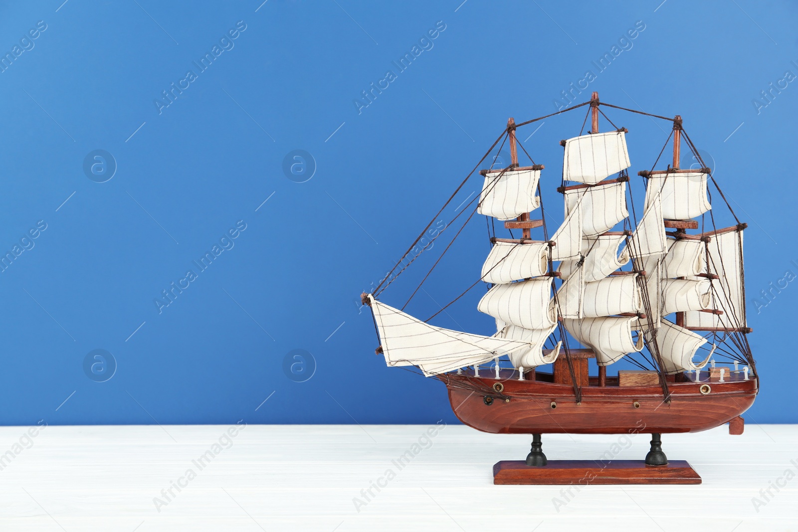Photo of Miniature model of old ship with white sails on table against blue background, space for text