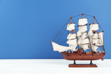 Photo of Miniature model of old ship with white sails on table against blue background, space for text