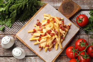 Photo of Delicious French fries with bacon, cheese sauce, tomatoes and herbs on wooden table, flat lay