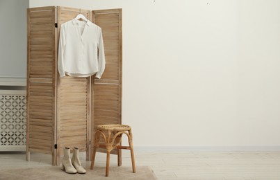 Stylish white shirt hanging on wooden folding screen, stool and boots indoors, space for text