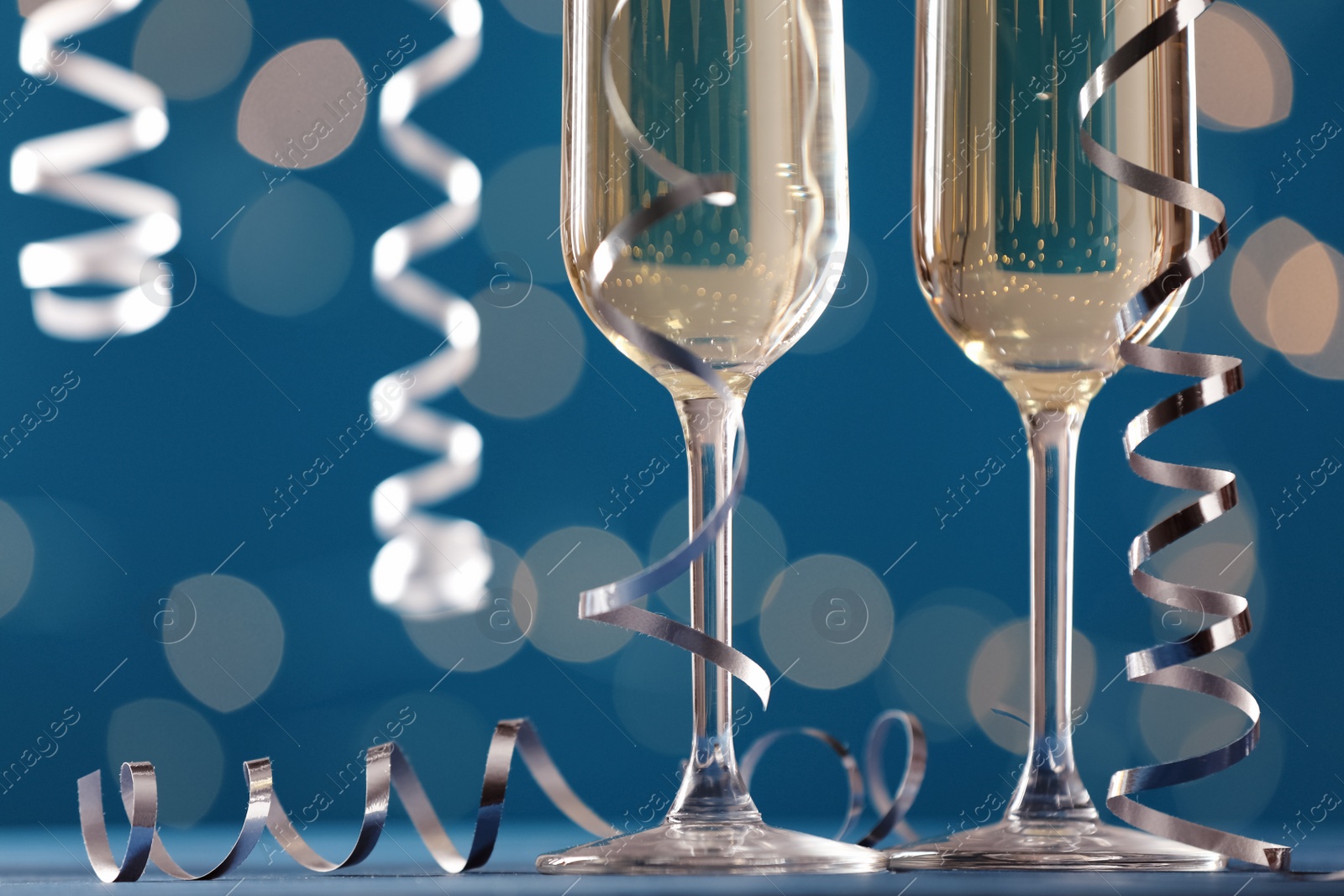 Photo of Glasses of champagne and serpentine streamers against blue background with blurred lights, closeup. Space for text