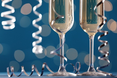 Photo of Glasses of champagne and serpentine streamers against blue background with blurred lights, closeup. Space for text