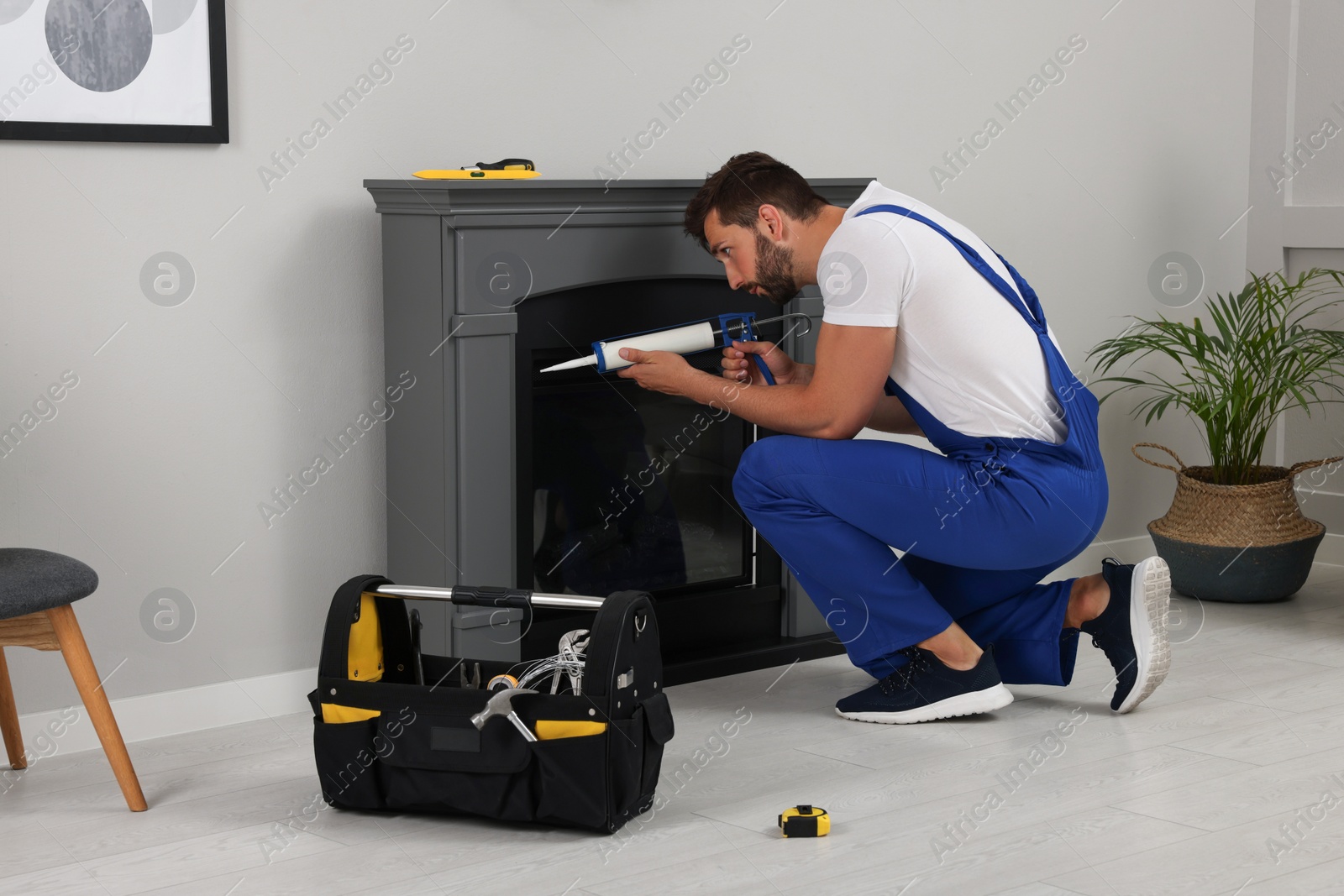 Photo of Professional technician sealing electric fireplace with caulk near white wall in room