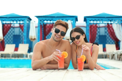 Photo of Woman in bikini and her boyfriend with refreshing cocktails near swimming pool on sunny day. Happy young couple