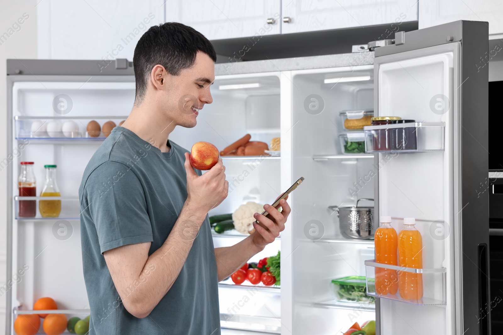 Photo of Happy man with apple and smartphone near refrigerator in kitchen