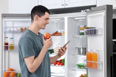 Happy man with apple and smartphone near refrigerator in kitchen