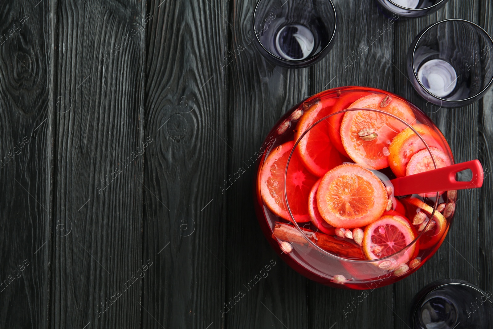 Photo of Bowl of delicious aromatic punch drink, ladle and empty glasses on black wooden table, flat lay. Space for text