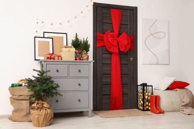 Photo of Beautiful fir tree and sack with Christmas gifts near chest of drawers and wooden door decorated with red bow in room