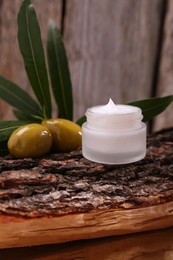 Photo of Glass jar of cream, olives and leaves on log near wooden wall, closeup