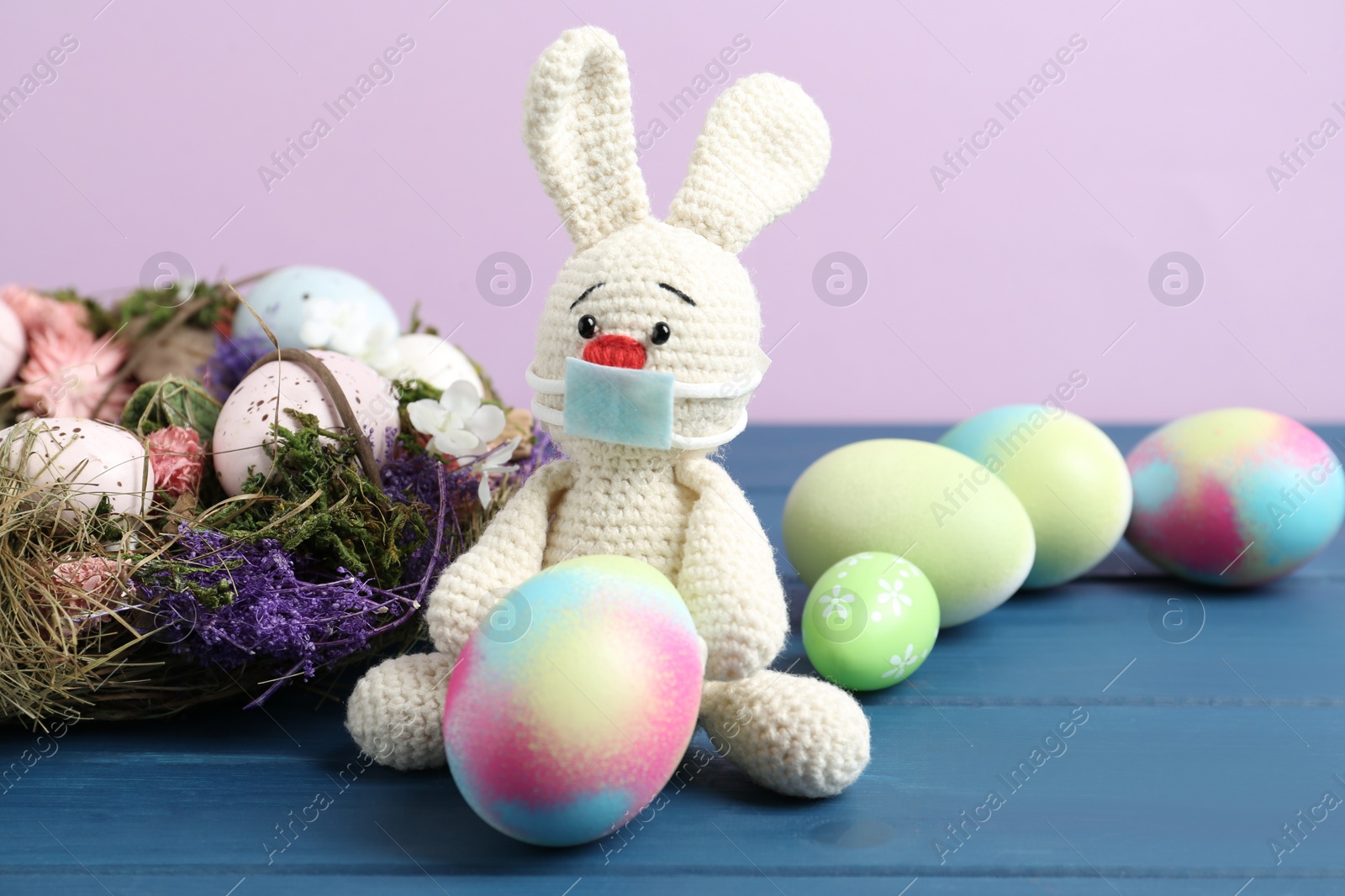 Photo of Toy bunny with protective mask and painted eggs on blue wooden table. Easter holiday during COVID-19 quarantine