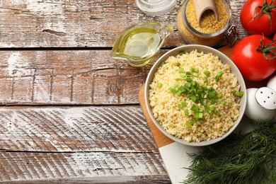 Photo of Delicious bulgur with green onion in bowl, tomatoes, dill and oil on wooden table, top view. Space for text