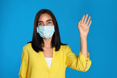 Photo of Young woman in protective mask showing hello gesture on light blue background. Keeping social distance during coronavirus pandemic