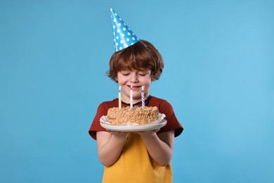 Photo of Birthday celebration. Cute little boy in party hat holding tasty cake with burning candles on light blue background