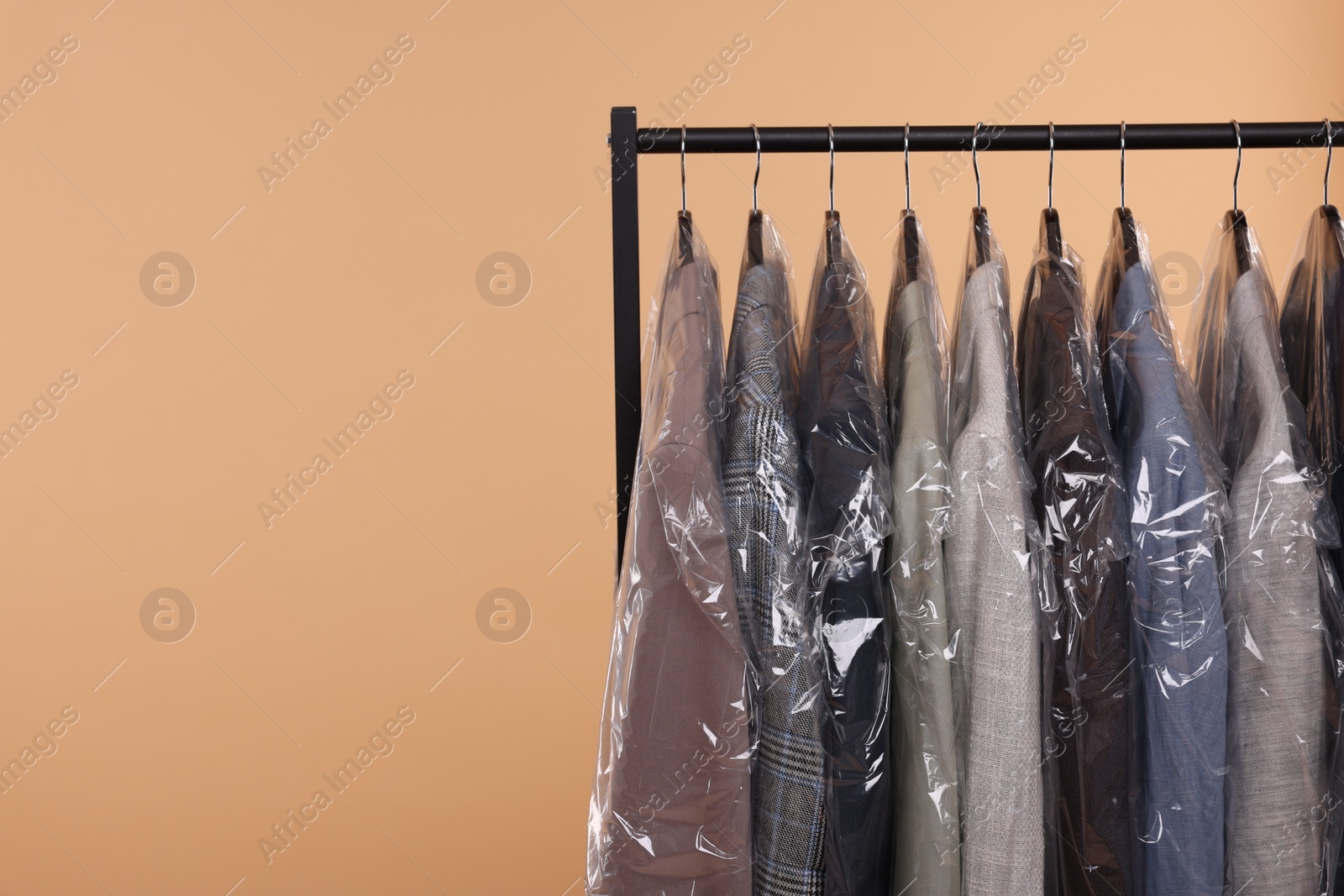 Photo of Dry-cleaning service. Many different clothes in plastic bags hanging on rack against beige background, space for text