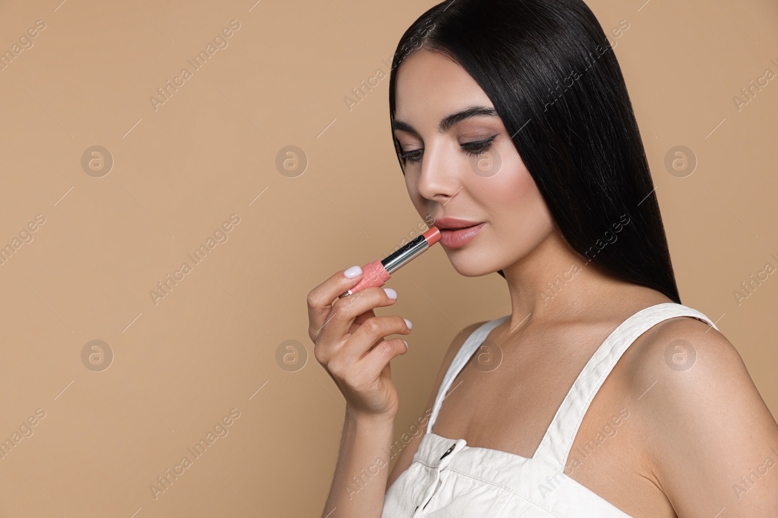 Photo of Beautiful young woman applying nude lipstick on beige background, space for text