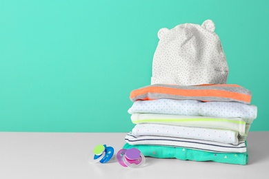 Stack of stylish child clothes and pacifiers on table against color background