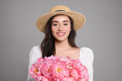 Photo of Beautiful young woman in straw hat with bouquet of pink peonies against grey background