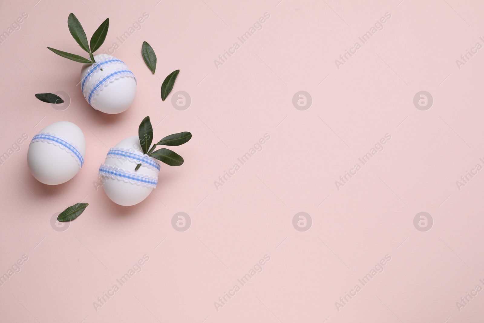 Photo of Beautifully decorated Easter eggs and green leaves on pale pink background, flat lay. Space for text
