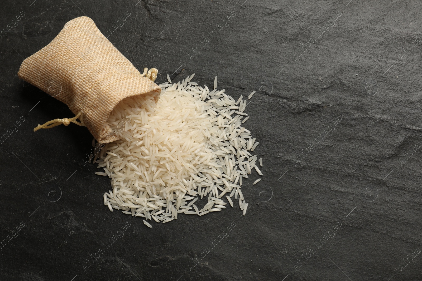 Photo of Raw basmati rice and overturned sack on black table, top view. Space for text