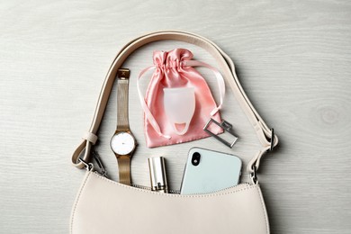 Menstrual cup and handbag with different women's accessories on white wooden table, flat lay