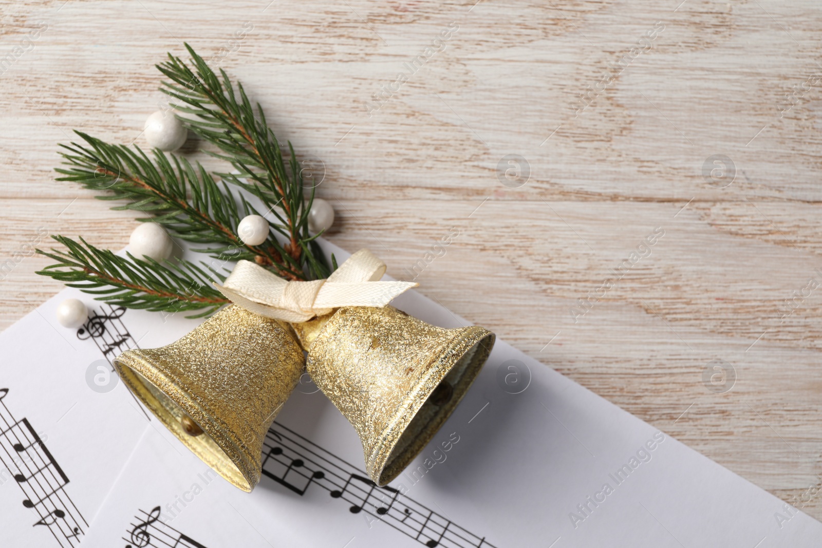 Photo of Golden shiny bells with bow, music sheets and fir twig on wooden table, flat lay. Space for text. Christmas decoration