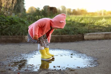Photo of Little girl wearing rubber boots standing in puddle outdoors, space for text. Autumn walk