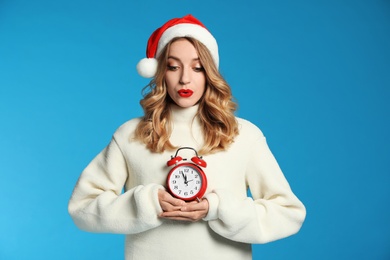 Photo of Happy young woman wearing Santa hat with alarm clock on light blue background. Christmas time