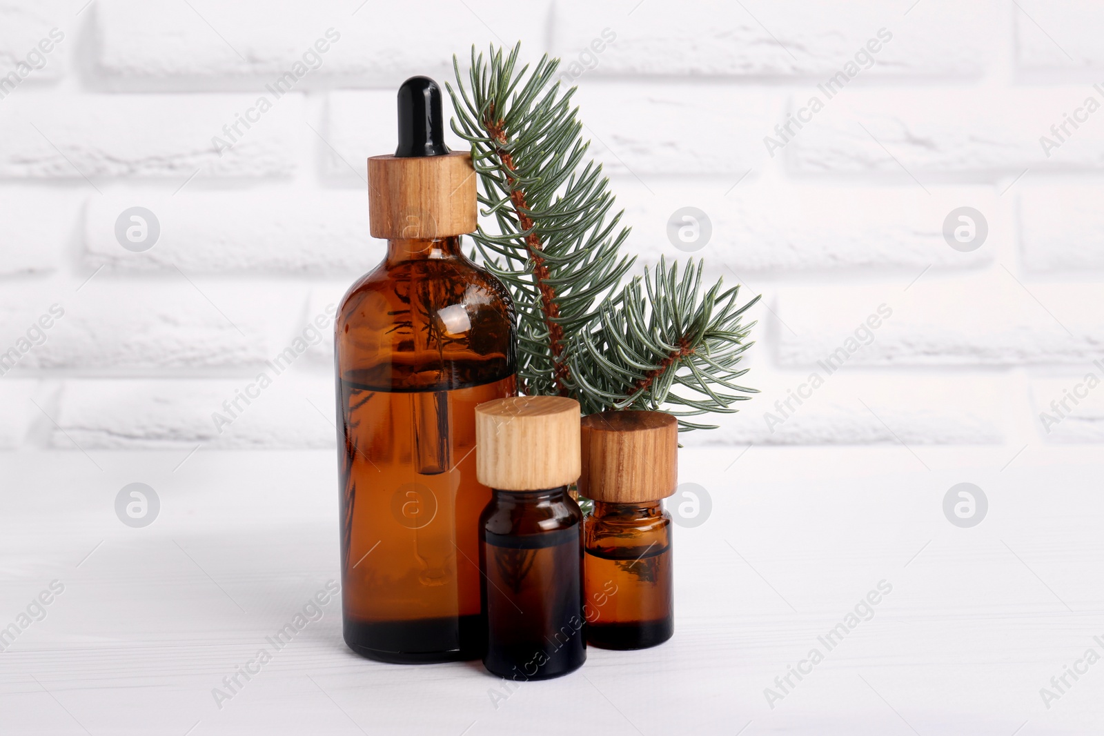 Photo of Bottles of essential oil and pine branch on white wooden table near brick wall