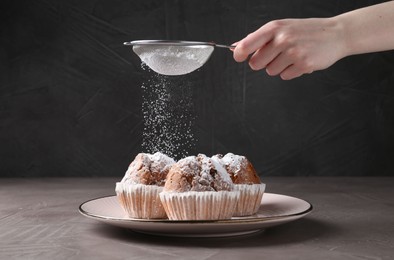 Photo of Woman with sieve sprinkling powdered sugar onto muffins at grey textured table, closeup