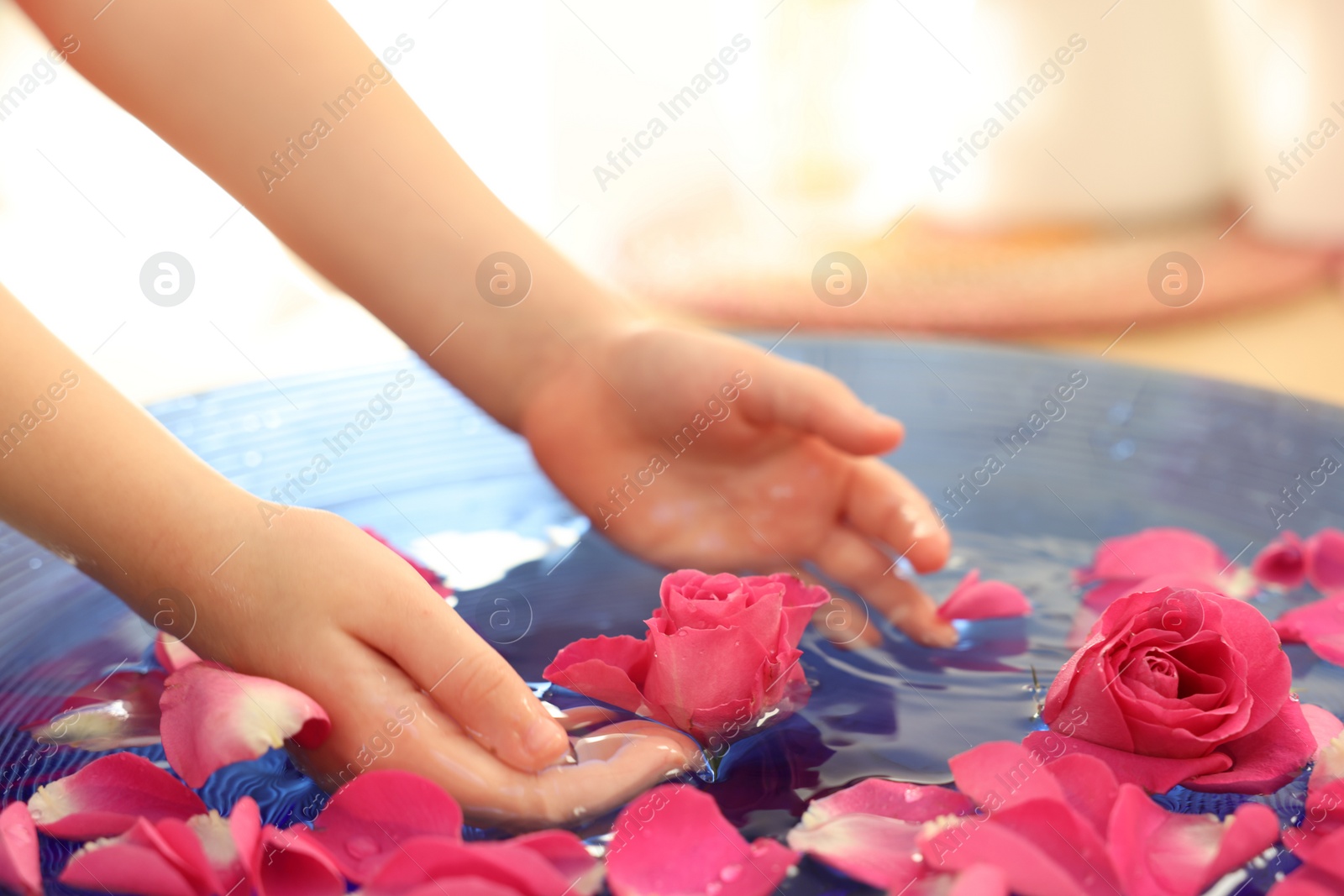 Photo of Child soaking hands in bowl with water and rose petals on blurred background, closeup