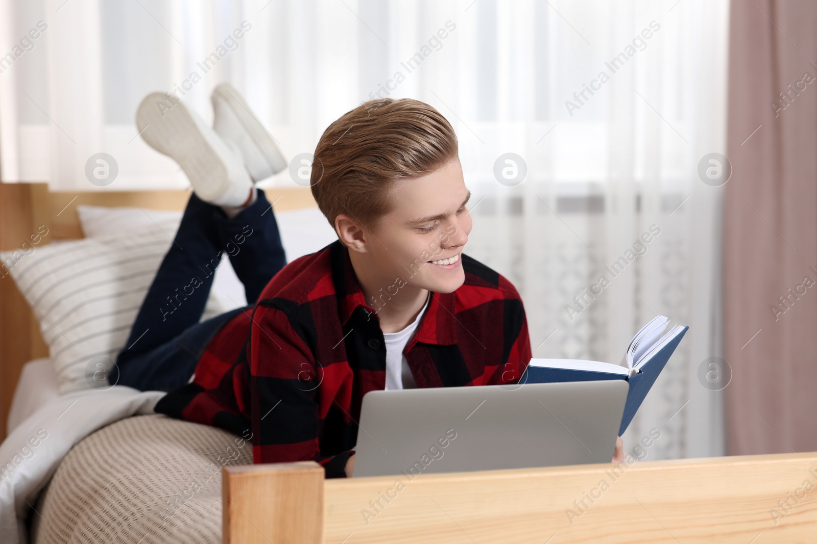 Photo of Online learning. Smiling teenage boy reading book near laptop on bed at home